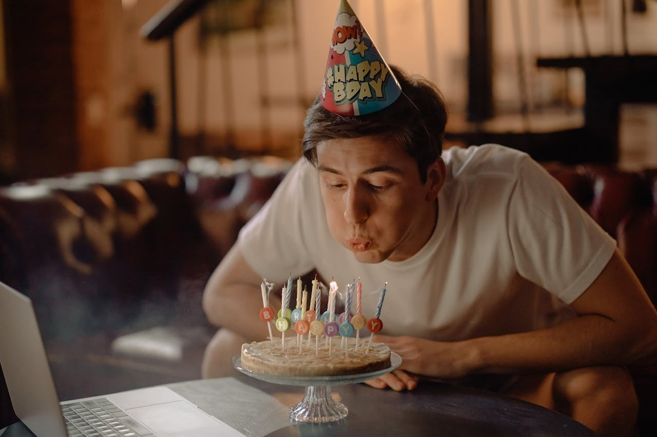 Person in a party hat blowing out birthday candles near a laptop.